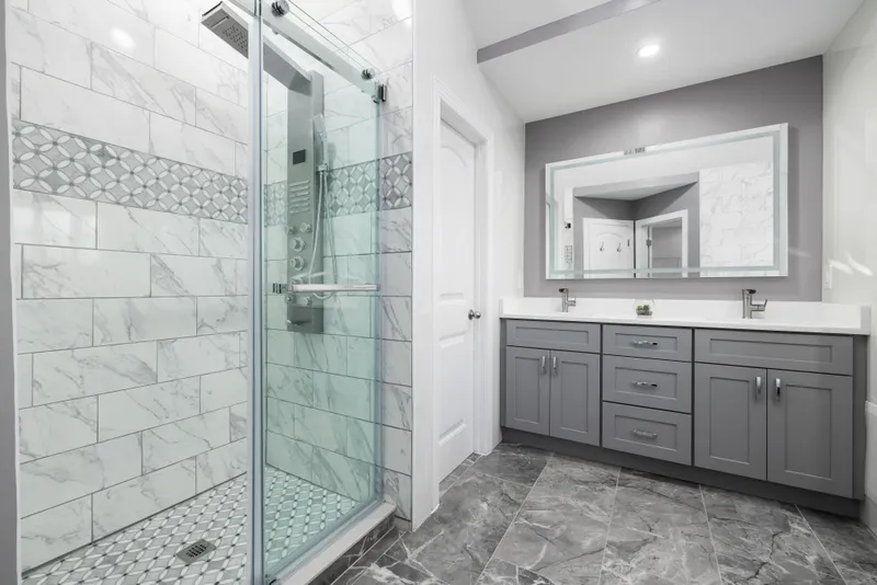 A finished bathroom combines a glass shower, patterned tile details, a gray double vanity, and a light countertop.