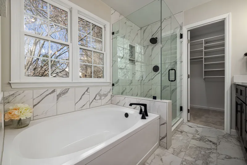 A tiled tub surround meets a glass shower with black fixtures beside a trimmed double window.