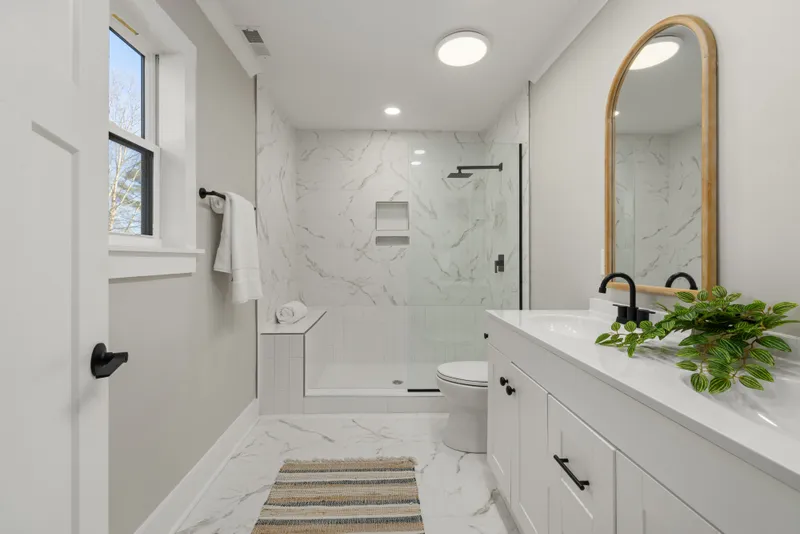Light veined wall and floor tile, a glass shower, and black fixtures complete a compact bathroom beside a white vanity.