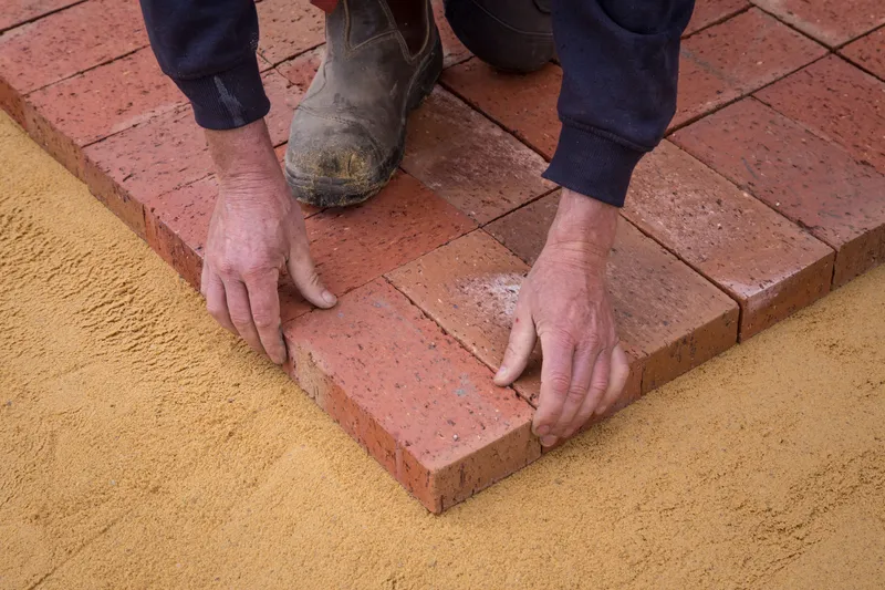 A worker aligns brick pavers on a prepared sand bed for a patio.
