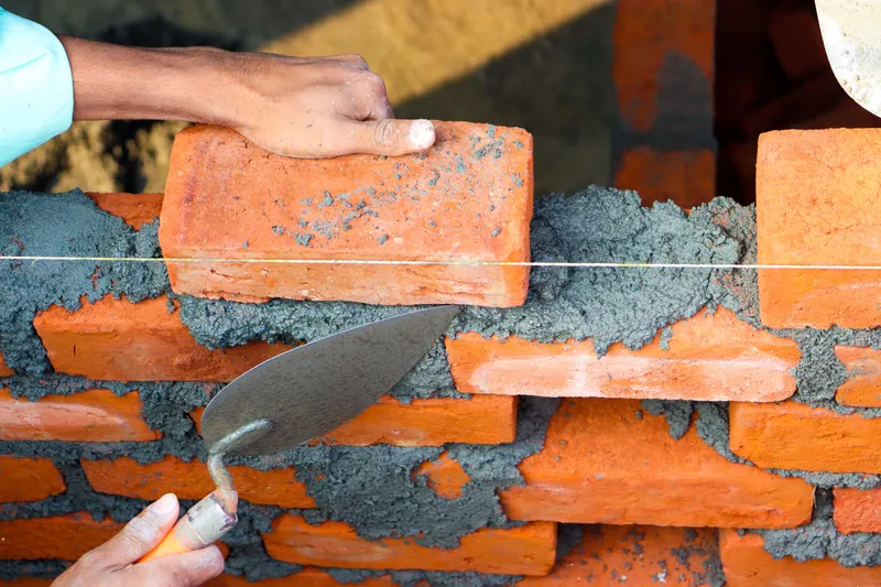 A mason places a brick along a guide line and spreads mortar with a trowel.