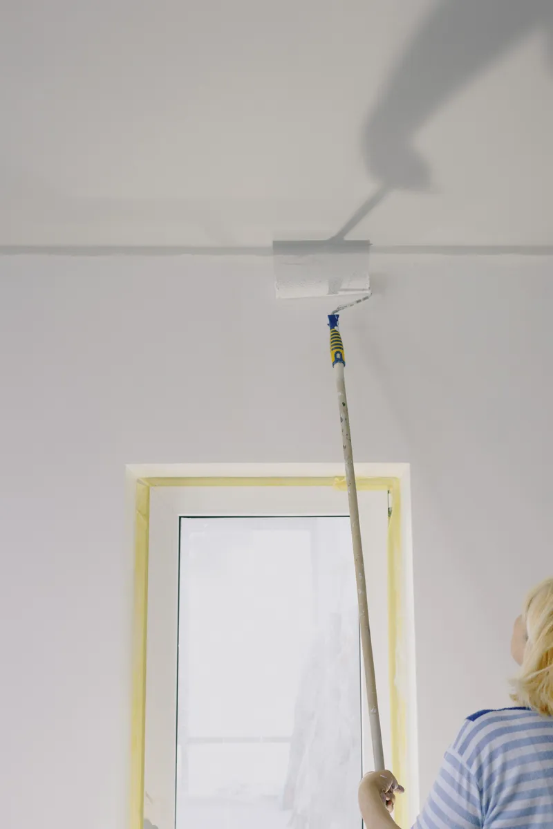 A worker uses an extension roller to paint a white ceiling in a room under renovation.
