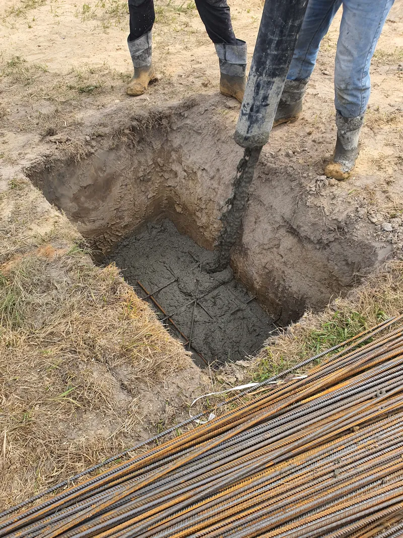A concrete pump hose fills an excavated foundation hole beside reinforcing steel and workers in work boots.