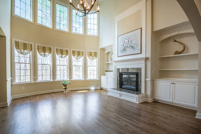 A double-height living room combines tall windows, wood flooring, built-in cabinets, and a detailed fireplace surround.
