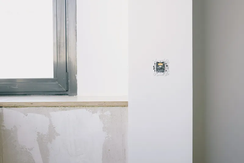 An open electrical box sits in an unfinished wall beside a window during an interior renovation.
