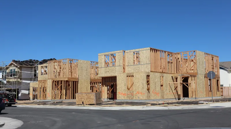 A two-story house under construction shows wood studs, OSB wall sheathing, and framed window openings beside a paved street.