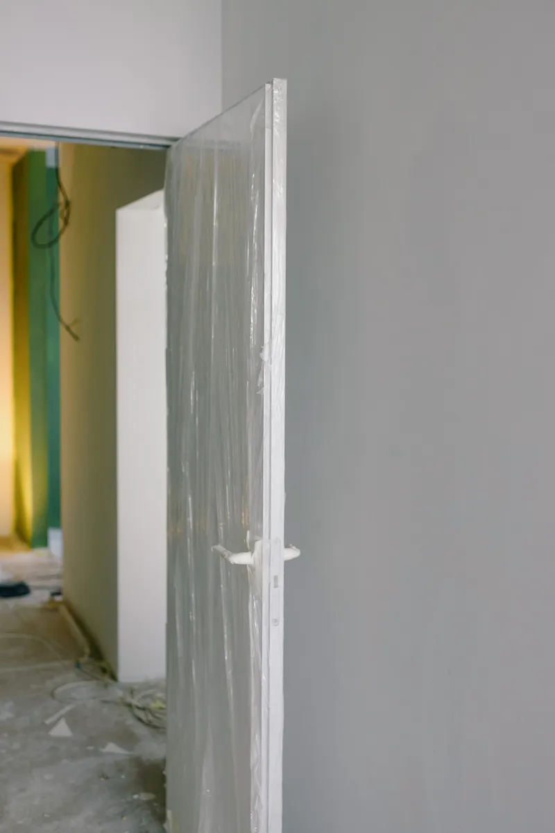 An installed interior door remains wrapped in protective plastic while adjacent rooms undergo finish work.