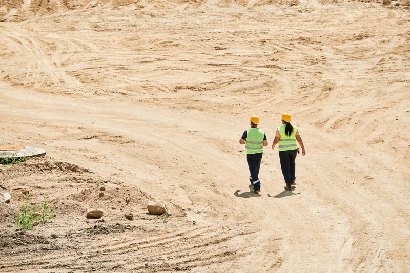 Two workers in hard hats and high-visibility vests walk across a graded construction site, viewed from behind.