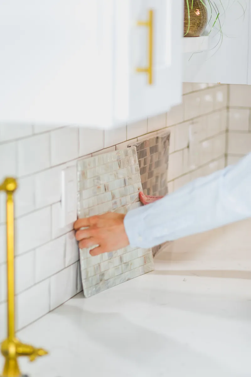 A hand holds a decorative tile sample against a kitchen backsplash beside a sink.