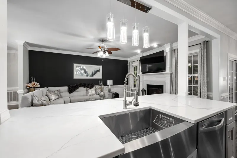 A finished kitchen island has a light veined countertop, deep apron-front sink, and pull-down faucet facing the living room.