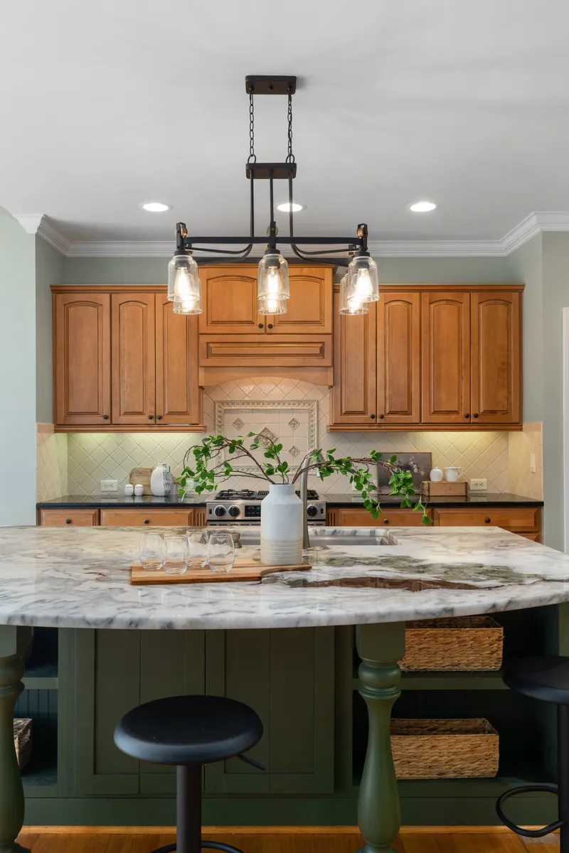 A kitchen island with a veined countertop sits before wood cabinets, a matching hood surround, and a patterned tile backsplash.