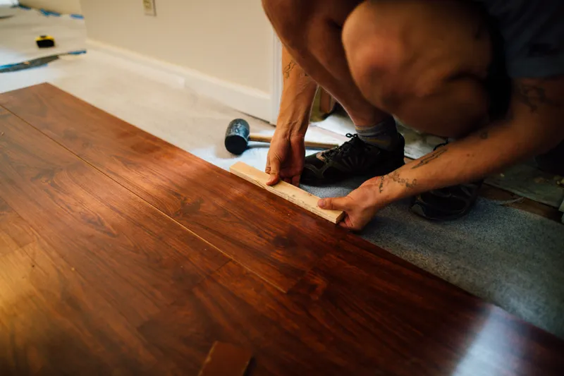 A flooring installer seats a laminate plank with a wood tapping block beside completed rows.