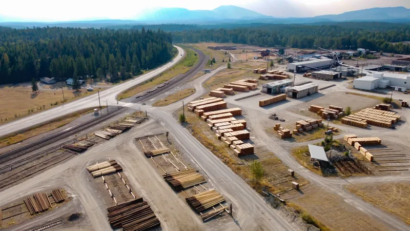 An aerial view shows lumber stacks, storage buildings, and vehicle access lanes at a lumber yard.