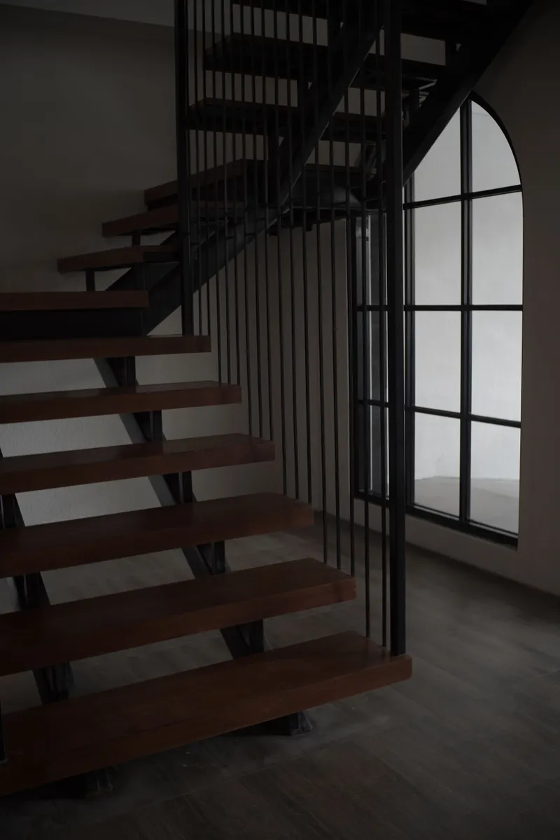 Open wood stair treads and dark metal railing frame a finished interior stair beside a tall window.