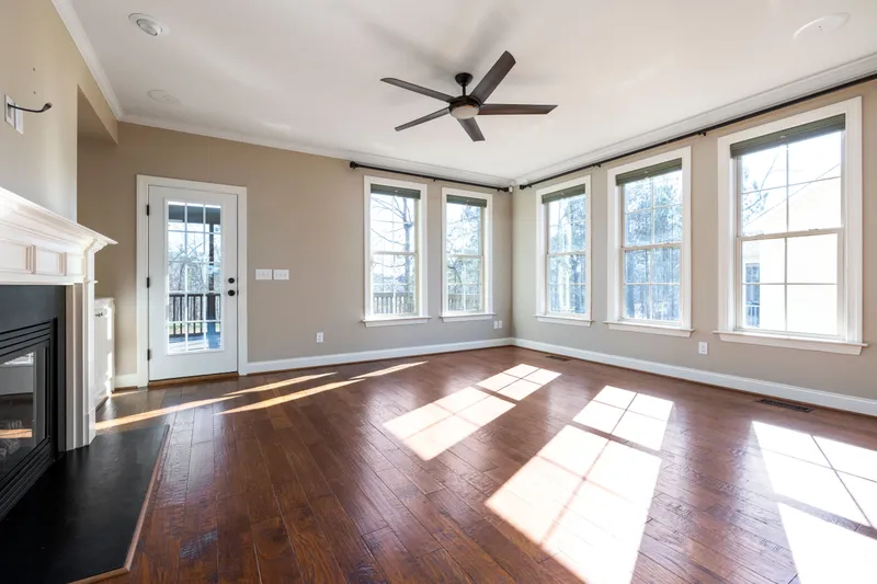 Sunlight falls across finished wood flooring beside tall trimmed windows, a glazed exterior door, and a fireplace.