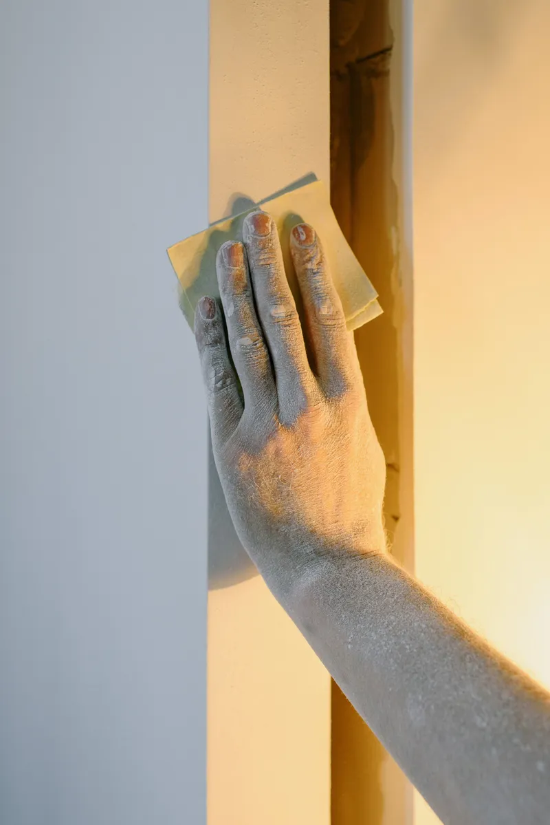 A worker sands an interior wall by hand beside a door opening before painting.