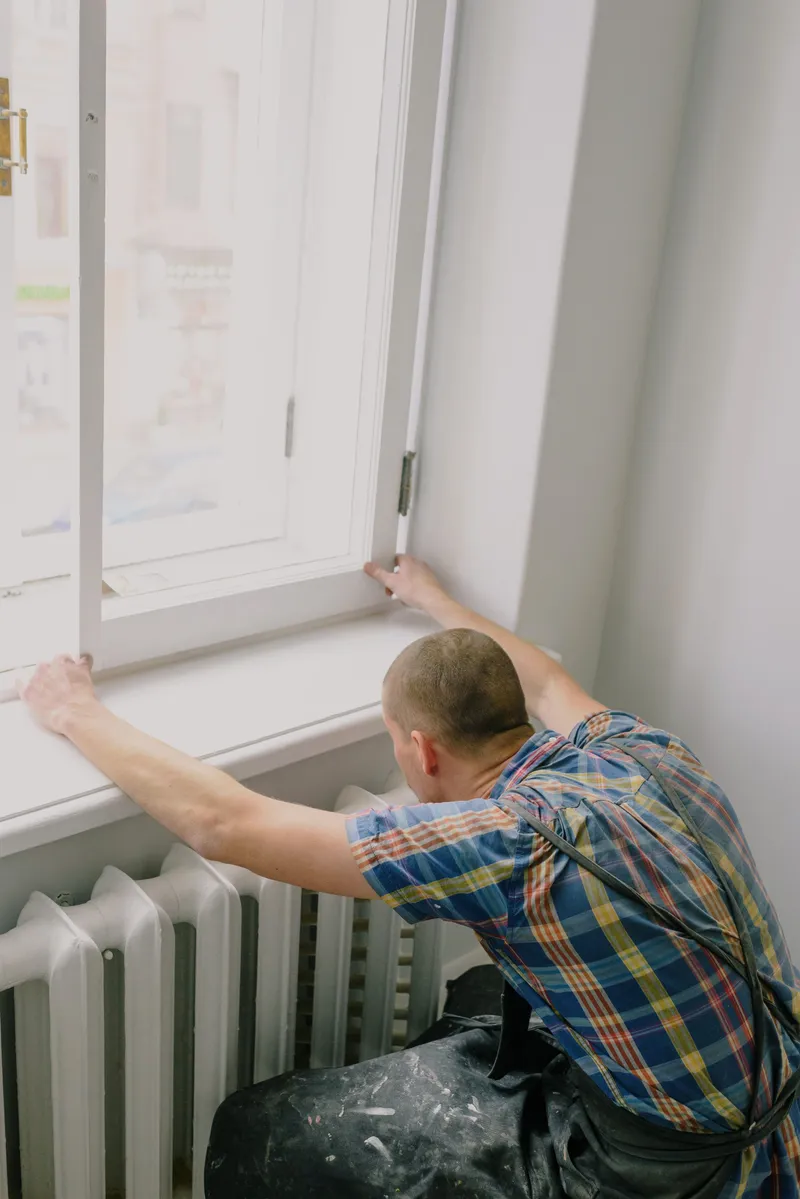 A worker viewed from behind adjusts an open window above a radiator during a home renovation.