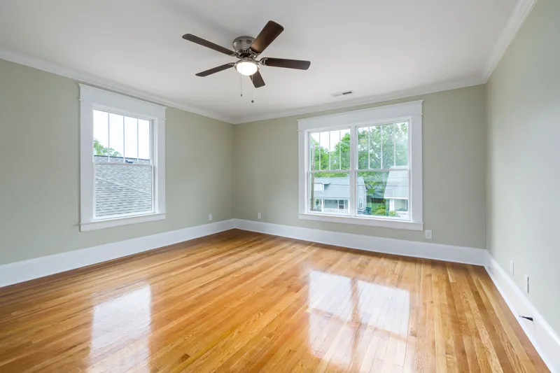 Sunlight crosses finished wood flooring beneath white window casing, baseboards, and crown molding in an empty room.